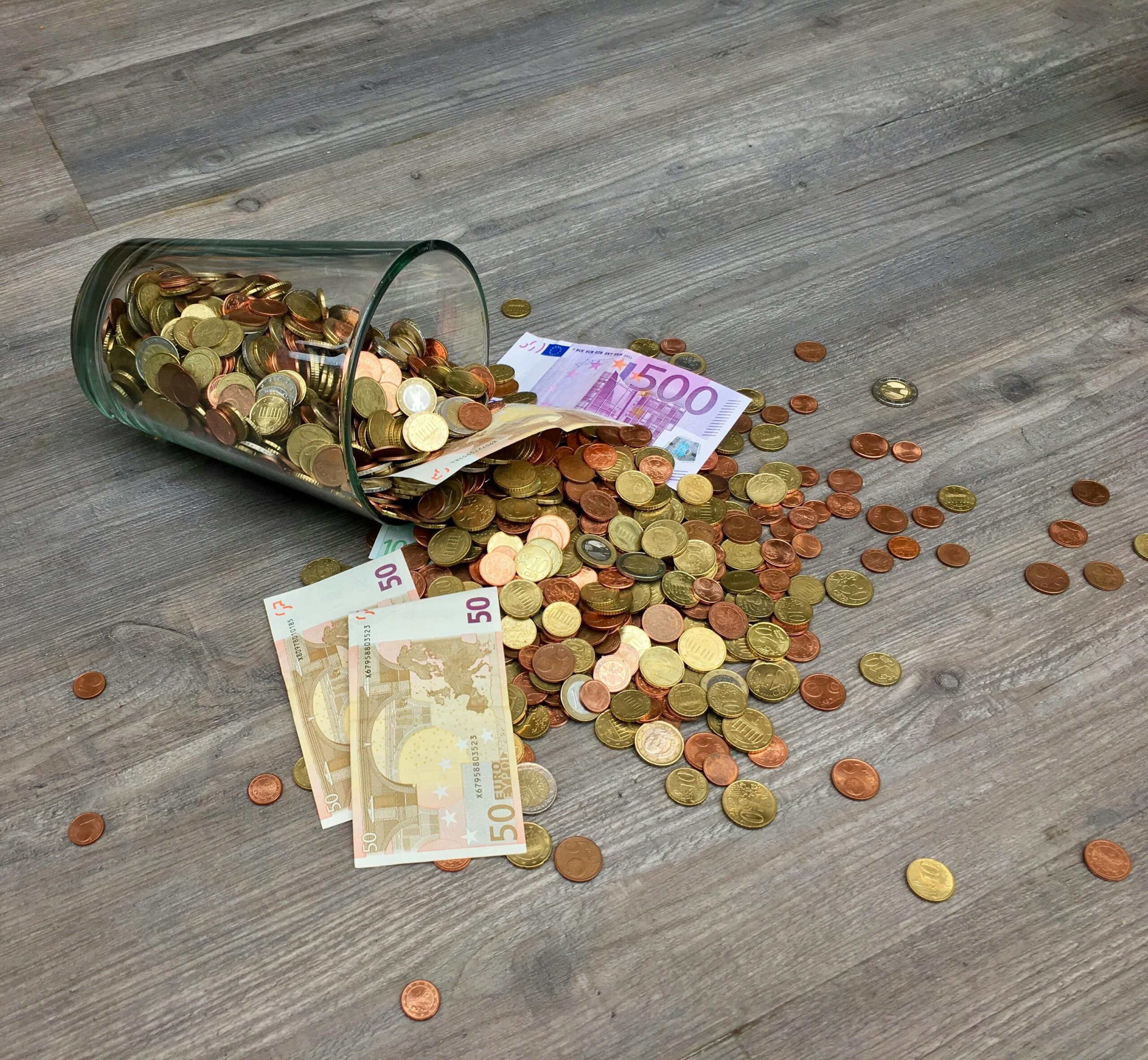 A glass jar with euro coins and banknotes spilled on a wooden surface, representing finance and savings.